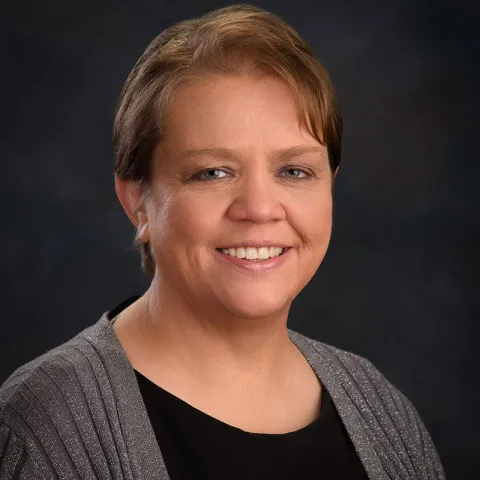 Professional headshot of a smiling woman with short brown hair, wearing a black top with a shimmering gray cardigan, set against a dark gradient background.