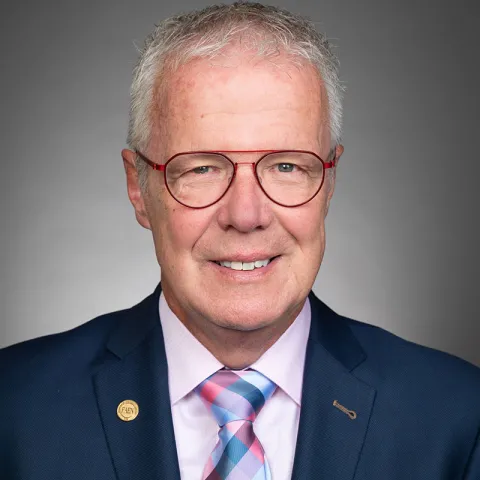 Professional headshot of a smiling man with short gray hair, wearing red-framed glasses, a navy blue suit, a light pink dress shirt, and a pink and blue plaid tie, set against a neutral gray background.