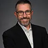 Headshot of a man with short dark hair, a beard, and glasses, wearing a black blazer over a white collared shirt, smiling confidently.