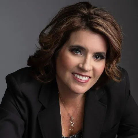 Professional headshot of a smiling woman with shoulder-length wavy brown hair, wearing a black blazer and a silver necklace, set against a dark gray background.