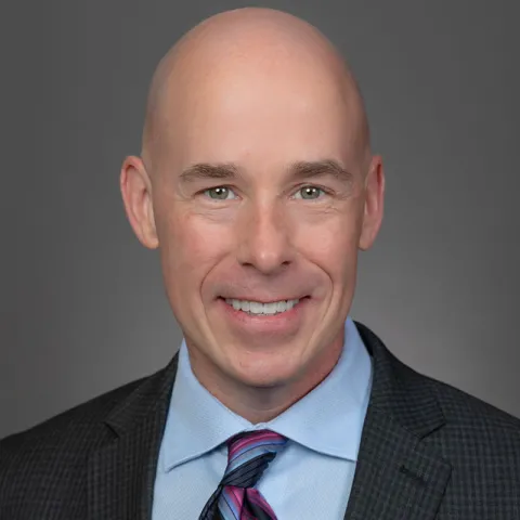 Professional headshot of a smiling bald man wearing a dark checkered suit, light blue dress shirt, and a pink and blue striped tie, set against a neutral gray background.