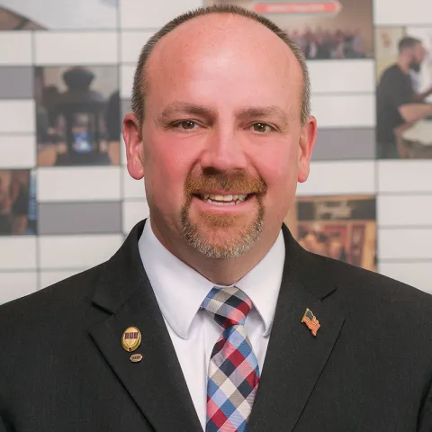 Professional headshot of a smiling man with short brown hair and a goatee, wearing a black suit, white shirt, and a red, white, and blue plaid tie, adorned with a U.S. flag lapel pin and a gold emblem, set against a background featuring a wall with photos and graphics.