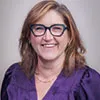 Headshot of a woman with shoulder-length light brown hair, wearing glasses, a purple blouse, and a necklace, smiling warmly.