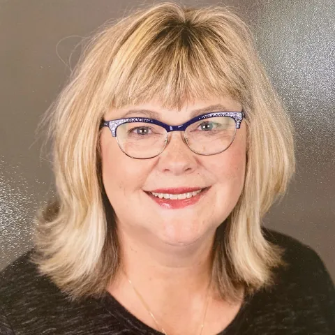 Professional headshot of a smiling woman with shoulder-length blonde hair, wearing blue-framed glasses and a black top, set against a neutral background.