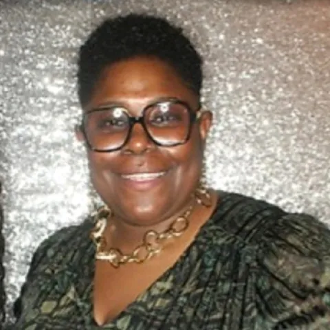 Headshot of a woman with short black hair, wearing large glasses, a patterned top, and a gold chain necklace, smiling warmly against a sparkling silver backdrop.