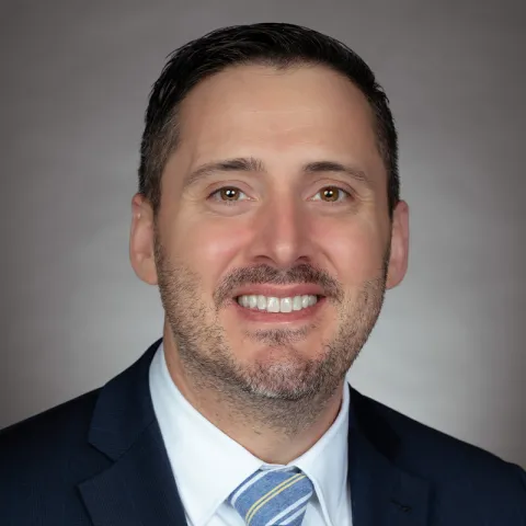 Professional headshot of a smiling man with short dark hair and light stubble, wearing a navy blue suit, white dress shirt, and a blue and yellow striped tie, set against a neutral gray background.