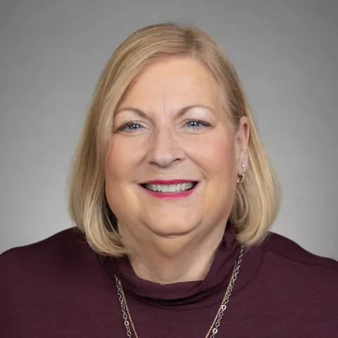 Professional headshot of a smiling woman with shoulder-length blonde hair, wearing a burgundy turtleneck sweater and a gold necklace, set against a neutral gray background.