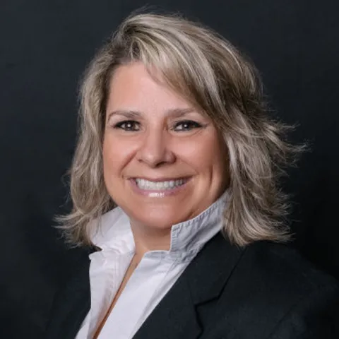 Professional headshot of a smiling woman with shoulder-length blonde hair, wearing a black blazer over a white collared shirt, set against a dark background.