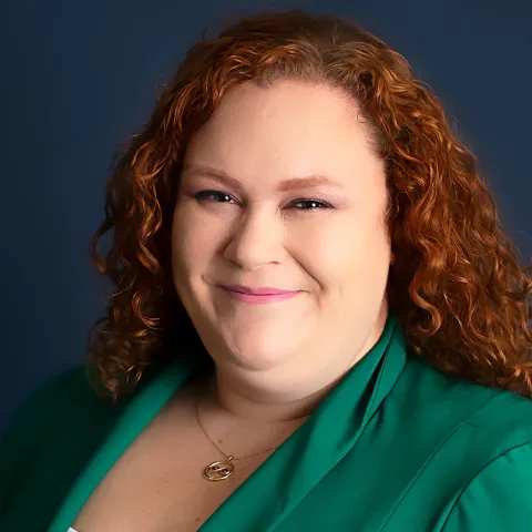 Professional headshot of a smiling woman with curly red hair, wearing a green blazer and a gold necklace, set against a dark blue background.