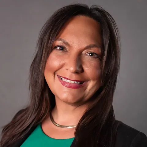 Professional headshot of a smiling woman with long dark brown hair, wearing a black blazer over a green top and a gold necklace, set against a neutral gray background.