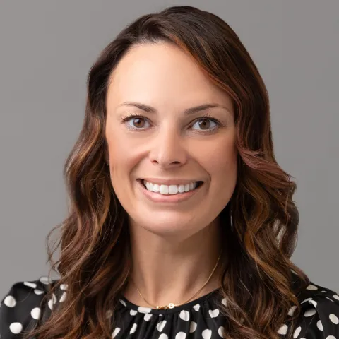 Professional headshot of a smiling woman with shoulder-length wavy brown hair, wearing a black and white polka dot blouse and a delicate gold necklace, set against a neutral gray background.