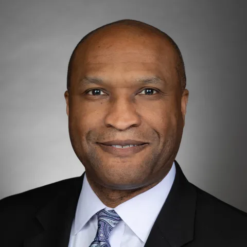 Professional headshot of a smiling man with a shaved head, wearing a black suit, white dress shirt, and a patterned tie, set against a neutral gray background.