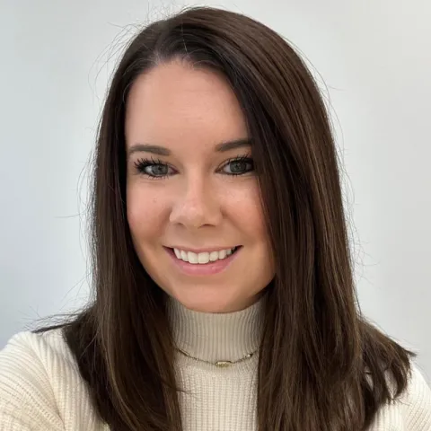 Professional headshot of a woman with straight brown hair, wearing a cream turtleneck sweater and a delicate gold necklace, smiling in front of a light gray background.