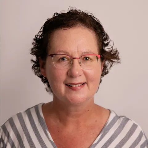 Headshot of a woman with short, curly brown hair wearing red-framed glasses and a striped top, smiling softly against a light neutral background.